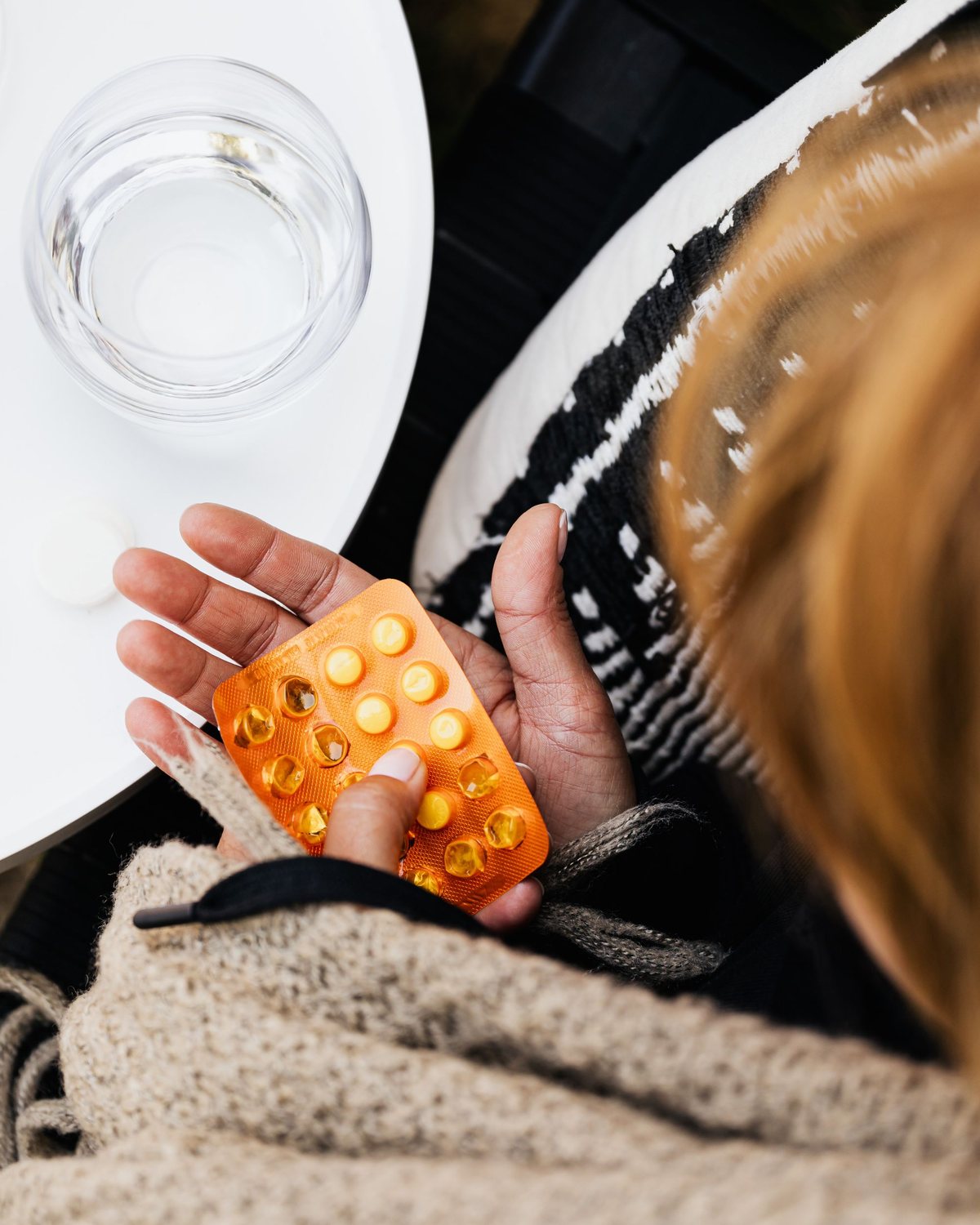 Vue de dessus : une main tient une plaquette de capsules orange, un verre d’eau posé sur une table blanche, un plaid beige